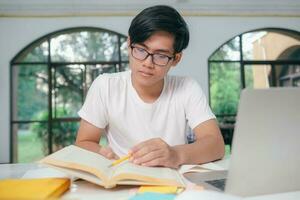 joven asiático masculino estudiante es preparando a leyendo un libros para exámenes a universidad. foto