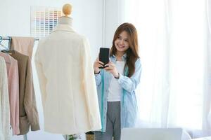joven asiático hembra Moda diseñador es tomando foto de su traje producto por utilizando un teléfono inteligente en un diseño estudio.