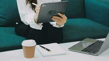 Happy Asian teen girl holding pad computer gadget using digital tablet technology sitting on the couch at home. Smiling young woman using apps, shopping online, reading news, browsing internet on sofa video