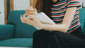 Happy Asian teen girl holding pad computer gadget using digital tablet technology sitting on the couch at home. Smiling young woman using apps, shopping online, reading news, browsing internet on sofa video