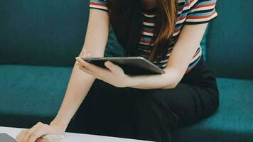 Happy Asian teen girl holding pad computer gadget using digital tablet technology sitting on the couch at home. Smiling young woman using apps, shopping online, reading news, browsing internet on sofa video