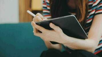 Happy Asian teen girl holding pad computer gadget using digital tablet technology sitting on the couch at home. Smiling young woman using apps, shopping online, reading news, browsing internet on sofa video