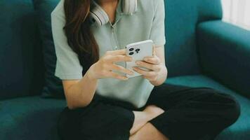 Happy Asian teen girl holding pad computer gadget using digital tablet technology sitting on the couch at home. Smiling young woman using apps, shopping online, reading news, browsing internet on sofa video