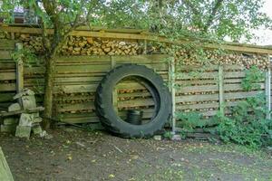 The wheel from the tractor is leaning against the woodpile. Woodpile behind the fence. There is vegetation, a tree grows. Lying bricks and stone blocks. photo