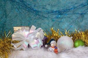 A smiling snowman on white fur next to Christmas balls and a large white bow. photo