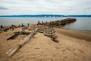 antiguo roto barco muelle en el isla de olkhon en lago Baikal. podrido barras y registros en el apuntalar de piedras y arena. detrás el montaña lago. el clima es sombrío, el cielo es gris. foto