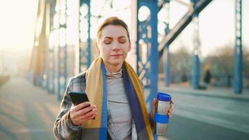 portret van een jong Kaukasisch zakenvrouw in een jas, wandelen aan de overkant de brug Aan een ijzig zonnig ochtend, drinken koffie en gebruik makend van smartphone. communicatie, werk dag, bezig leven concept video