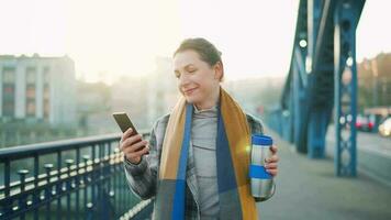 Portrait of a young caucasian businesswoman in a coat, walking across the bridge on a frosty sunny morning, drinking coffee and using smartphone. Communication, work day, busy life concept video