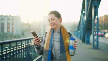 portret van een jong Kaukasisch zakenvrouw in een jas, wandelen aan de overkant de brug Aan een ijzig zonnig ochtend, drinken koffie en gebruik makend van smartphone. communicatie, werk dag, bezig leven concept video