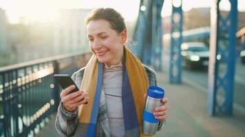 Porträt von ein jung kaukasisch Geschäftsfrau im ein Mantel, Gehen über das Brücke auf ein eisig sonnig Morgen, Trinken Kaffee und mit Smartphone. Kommunikation, Arbeit Tag, beschäftigt Leben Konzept video