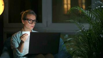 Woman is sitting in the armchair and makes an online purchase using a credit card and laptop at night video