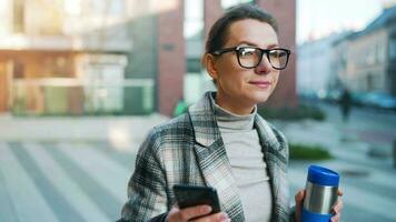 portret van een jong Kaukasisch zakenvrouw met bril en een jas wandelingen door de bedrijf wijk, drinken koffie en gebruik makend van smartphone video