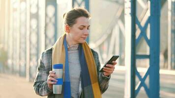 portret van een jong Kaukasisch zakenvrouw in een jas, wandelen aan de overkant de brug Aan een ijzig zonnig ochtend, drinken koffie en gebruik makend van smartphone. communicatie, werk dag, bezig leven concept video