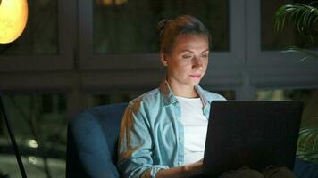 Woman is sitting in the armchair and makes an online purchase using a credit card and laptop at night video