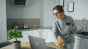 Woman with smartphone in hand standing in home cozy kitchen use laptop do freelance work, looking focused deep lost in thoughts video