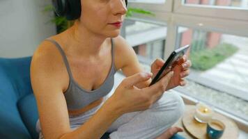 contento relajado mujer en inalámbrico auriculares escuchando a música y utilizando móvil aplicaciones o comunica en social redes en teléfono inteligente video