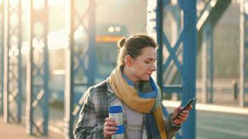 portret van een jong Kaukasisch zakenvrouw in een jas, wandelen aan de overkant de brug Aan een ijzig zonnig ochtend, drinken koffie en gebruik makend van smartphone. communicatie, werk dag, bezig leven concept video