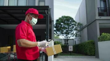 een levering Mens vervelend een rood overhemd wandelingen Holding een pakket doos en gebruik makend van zijn telefoon naar zoeken voor het naar de klanten huis. video