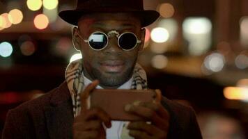 jong zwart Mens in wijnoogst stijl is gebruik makend van een slim telefoon in de stad straten Bij nacht video