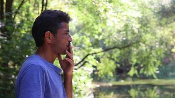 A young man with a mustache and beard smoking a cigarette while standing in the forest, an unhealthy smoking man in greenery video