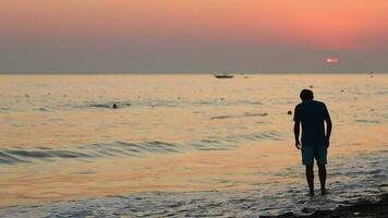 silhouette de une triste déprimé homme sur le plage pendant coucher de soleil, une vue de le Montagne et gens nager dans le mer dans le soir video