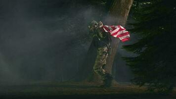 adulto maschio soldato nel pieno uniforme nel boschi a notte con americano bandiera librarsi al di sopra di il suo le spalle. video
