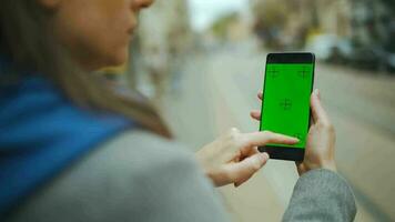 Woman at the street using smartphone with green mock-up screen in vertical mode against the backdrop of a passing tram video