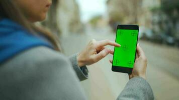 Woman at the street using smartphone with green mock-up screen in vertical mode against the backdrop of a passing tram video