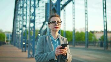 caucásico mujer de negocios en un Saco caminando alrededor el ciudad en el temprano Mañana y utilizando teléfono inteligente comunicación, trabajo día, ocupado vida concepto video