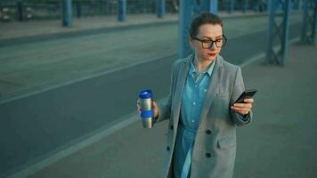 femme dans une manteau, en marchant autour le ville dans le de bonne heure matin, en buvant café et en utilisant téléphone intelligent, lent mouvement video