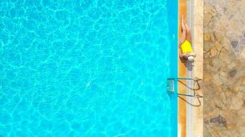 Aerial view of a woman in yellow swimsuit and a hat sits on the edge of the pool video