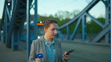 mujer en un abrigo, caminando alrededor el ciudad en el temprano mañana, Bebiendo café y utilizando teléfono inteligente, lento movimiento video