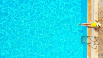 Aerial view of a woman in yellow swimsuit and a hat sits on the edge of the pool video