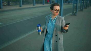 Woman in a coat, walking around the city in the early morning, drinking coffee and using smartphone, slow motion video