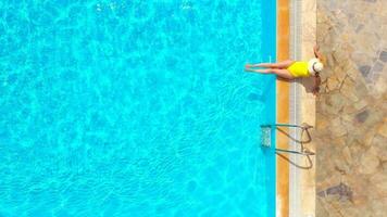 Aerial view of a woman in yellow swimsuit and a hat sits on the edge of the pool video