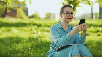 Busy attractive woman working at the laptop and using smartphone while sitting on grass in city park at sunset video