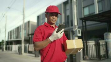 A parcel delivery worker wearing a red uniform is holding a parcel box to the recipient. At that time, he was using a mobile phone. contact the receiver in front of the house video