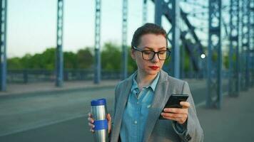 vrouw in een jas, wandelen in de omgeving van de stad in de vroeg ochtend, drinken koffie en gebruik makend van smartphone, langzaam beweging video