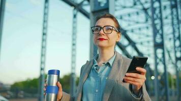 mujer en un abrigo, caminando alrededor el ciudad en el temprano mañana, Bebiendo café y utilizando teléfono inteligente, lento movimiento video