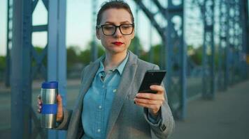 mujer en un abrigo, caminando alrededor el ciudad en el temprano mañana, Bebiendo café y utilizando teléfono inteligente, lento movimiento video