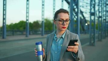 vrouw in een jas, wandelen in de omgeving van de stad in de vroeg ochtend, drinken koffie en gebruik makend van smartphone, langzaam beweging video