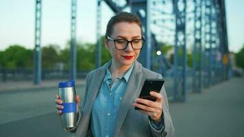 mujer en un abrigo, caminando alrededor el ciudad en el temprano mañana, Bebiendo café y utilizando teléfono inteligente, lento movimiento video