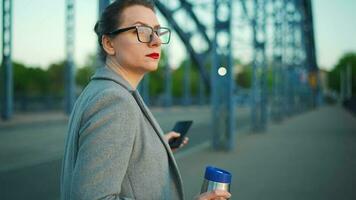 mujer en un abrigo, caminando alrededor el ciudad en el temprano mañana, Bebiendo café y utilizando teléfono inteligente, lento movimiento video