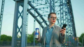 vrouw in een jas, wandelen in de omgeving van de stad in de vroeg ochtend, drinken koffie en gebruik makend van smartphone, langzaam beweging video