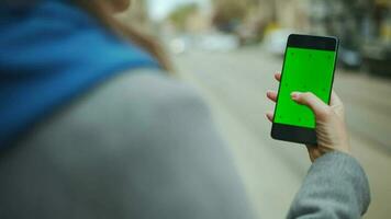 Woman at the street using smartphone with green mock-up screen in vertical mode against the backdrop of a passing tram video