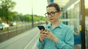 vrouw staat Bij een vervoer hou op, gebruik makend van smartphone en aan het wachten voor de tram video