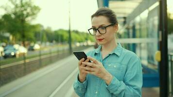 vrouw staat Bij een vervoer hou op, gebruik makend van smartphone en aan het wachten voor de tram video