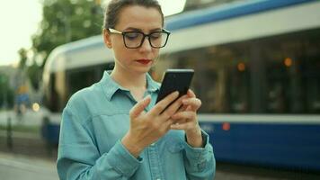 Frau steht beim ein Öffentlichkeit Transport halt und mit Smartphone. Straßenbahn zieht oben im das Hintergrund. schleppend Bewegung video