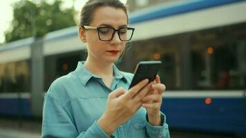 Frau steht beim ein Öffentlichkeit Transport halt und mit Smartphone. Straßenbahn zieht oben im das Hintergrund. schleppend Bewegung video