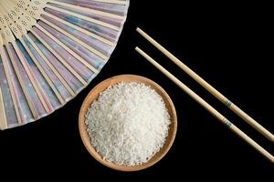 Raw white rice, hand fan and chopsticks on black background. Long uncooked rice in a wooden plate. photo